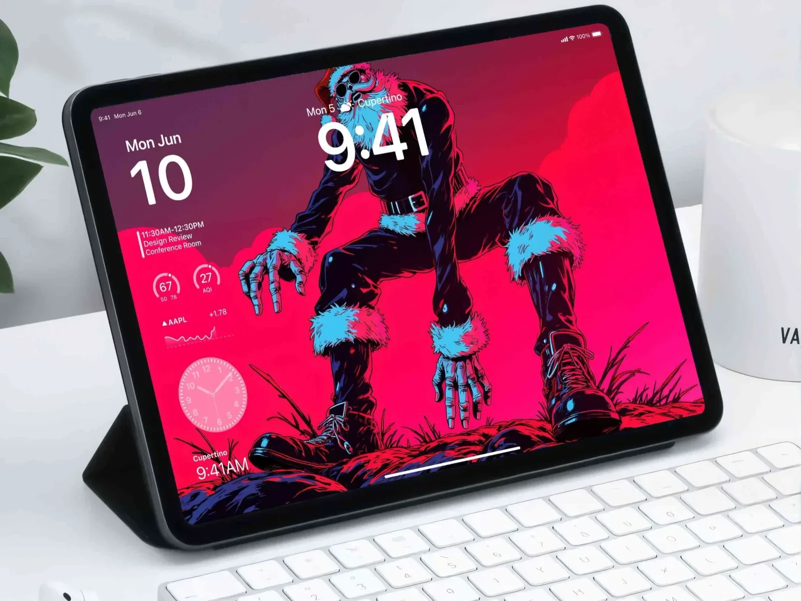 A tablet on a keyboard shows a comic Santa in a black suit with cyan fur, crouched on magenta rocks, with calendar and weather widgets along the left edge.